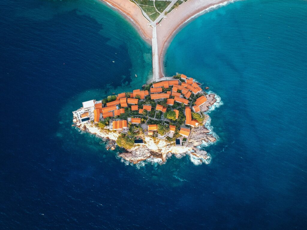 Sveti Stefan in Montenegro, a tiny island village with red-roofed buildings linked to the coast by a narrow strip of land, surrounded by bright blue sea.